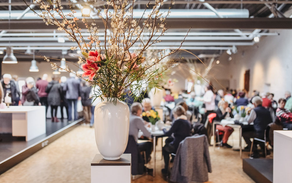 Porzellanvase mit großesm Blumenstrauß im Vordergrund, Personen an Tischen sitzend im Hintergrund